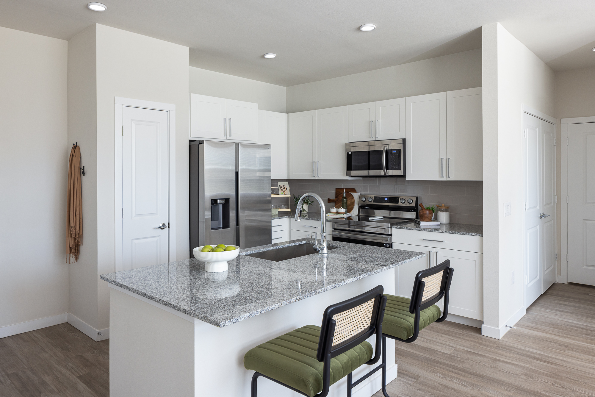 Modern kitchen with white cabinets, stainless steel appliances, granite countertops, and two green chairs at the island. A bowl of apples sits on the countertop. Prose Thunder Rock Apartments in Marble Fall, Texas.